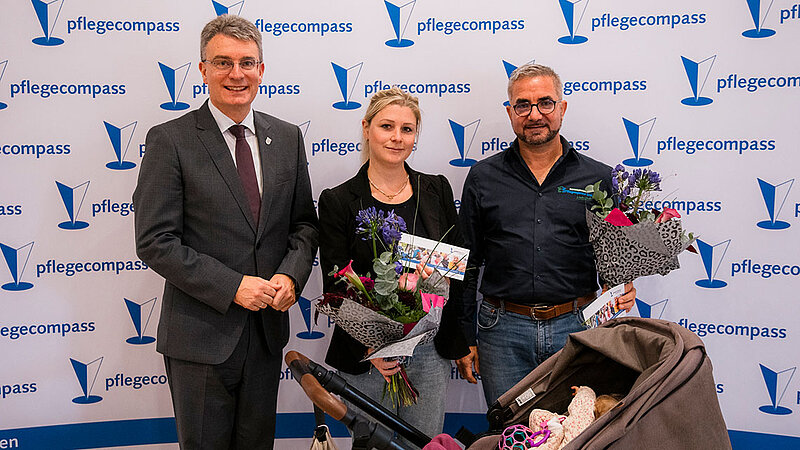Der Landrat und zwei Preisträger stehen zusammen. Die Preisträger halten Blumensträuße in den Händen. Ein Baby im Kinderwagen ist ebenfalls zu sehen.
