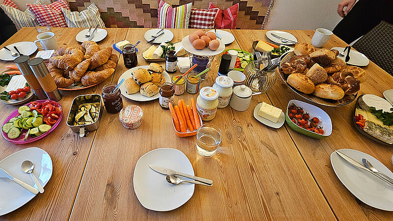 Ein reich gedeckter Frühstückstisch mit Brötchen, Aufstrich, Gemüse und Obst. 