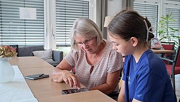 Eine Jugendliche und eine ältere Frau sitzen zusammen am Tisch. Vor ihnen liegt ein Handy.