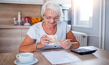 Seniorin sitzt mit Taschenrechner und Geldscheinen an einem Küchentisch.