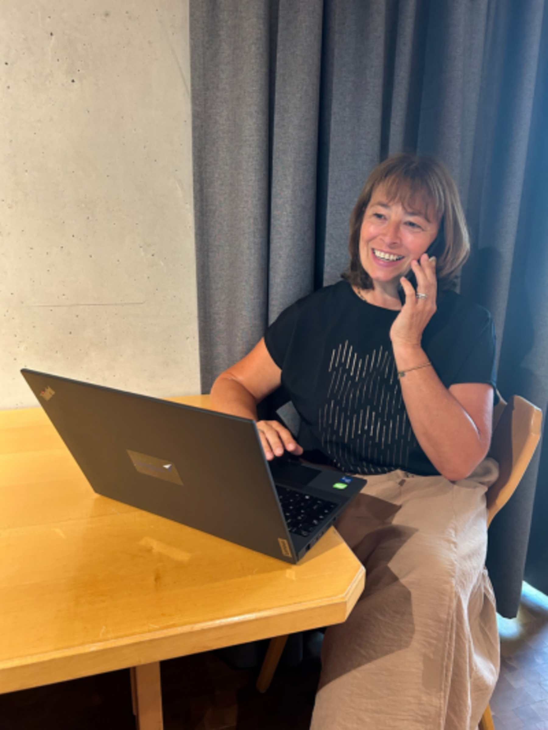 Eine Frau sitzt an einem Tisch. Der Laptop steht aufgeklappt vor ihr, sie telefoniert und lächelt.