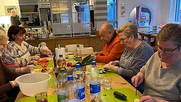 Gäste im Café Vergissmeinnicht bereiten Gemeinsam Essen am Tisch vor. Im Hintergrund ist eine Küche zu sehen.