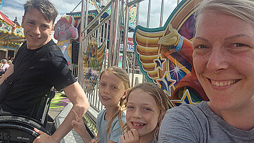 Ein erwachsener Mann im Rollstuhl, zwei Kinder und eine erwachsene Frau lachen in die Kamera. Im Hintergrund sieht man eine Kirmes.