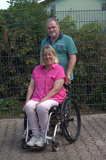 Auf dem Bild sind Rainer Seepe und Marion Kuehn-Seepe zu sehen. Rainer Seepe steht hinter seiner Frau und schiebt ihren Rollstuhl.