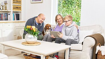 Eine Familie mit Oma, Vater, Kind und Hund sitzen auf dem Sofa und schauen gemeinsam auf ein Tablet.