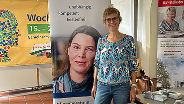 Frau steht vor einem Banner und lächelt in die Kamera. Vor ihr liegen Prospekte auf einem Tisch.
