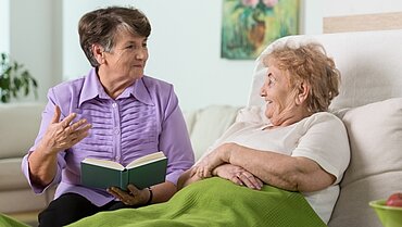 Eine ältere Frau liest einer anderen älteren Frau vor, die auf dem Sofa mit einer Decke sitzt.