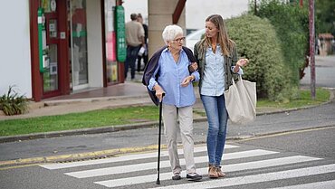 Eine jüngere Frau mit einer Stofftasche und eine ältere Frau mit einem Gehstock überqueren einen Zebrastreifen. Die jüngere Frau ist bei der älteren am Arm eingehakt.