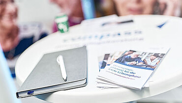 Infomaterial liegt auf einem Tisch mit compass-Logo. Im Hintergrund Roll-Ups.
