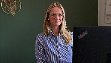 Nicole Zahn sitzt hinter ihrem Schreibtisch vor einer grünen Wand und lächelt in die Kamera. 