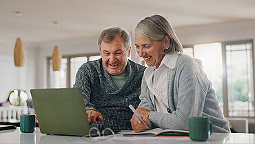 Ein Mann und eine Frau, die beide älter sind, schauen gemeinsam auf einen Computer. Sie lächeln und machen sich Notizen.