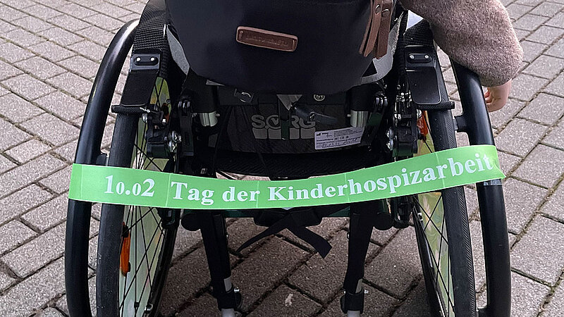 Am Rollstuhl eines Kindes ist ein grünes Band vom Tag der Kinderhospizarbeit befestigt. 