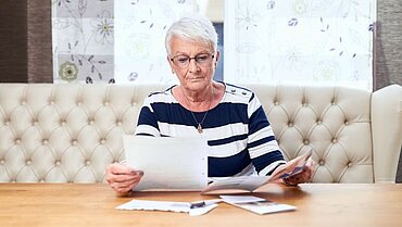 Seniorin mit kurzen Haaren sitzt an Tisch vor einem Fenster und sieht Dokumente durch.
