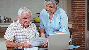 Ein Mann und eine Frau befinden sich vor einem Laptop und schauen auf einen Zettel.