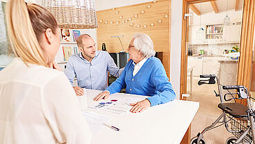 Das Bild zeigt einen älteren Herrn, seinen jüngeren Angehörigen und eine Pflegeberaterin. Sie sitzen am Küchentisch und sind in ein Gespräch vertieft.
