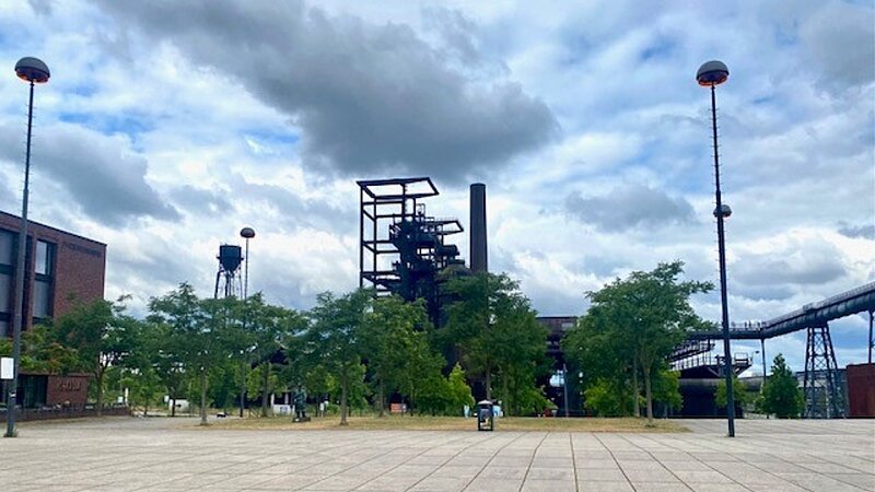 Ein Platz mit Bäumen und Stahlkonstruktionen vor bewölktem Himmel.  