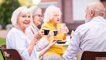 Vier ältere Menschen, drei Frauen und ein Mann, sitzen zusammen und trinken Kaffee. Eine der Frauen guckt in die Kamera und lächelt.
