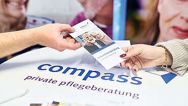 Ein Flyer für Präventionsberatung wird über einem Tisch mit compass-Logo von einer Hand in eine andere weitergereicht.