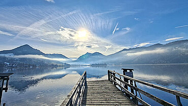 Auf dem Bild ist ein Steg zu sehen, der in einen großen See hinein führt. In ihm spiegeln sich die Berge am Horizont sowie der blaue Himmel und wenige kleine, weiße Wolken. Es handelt sich um den Schliersee in Bayern.