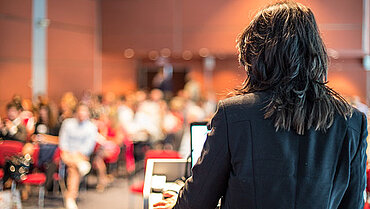 Eine Frau steht an einem Podium, den Rücken zur Kamera gewandt. Im Hintergrund sitzen Menschen zu ihr gewandt auf Stühlen.