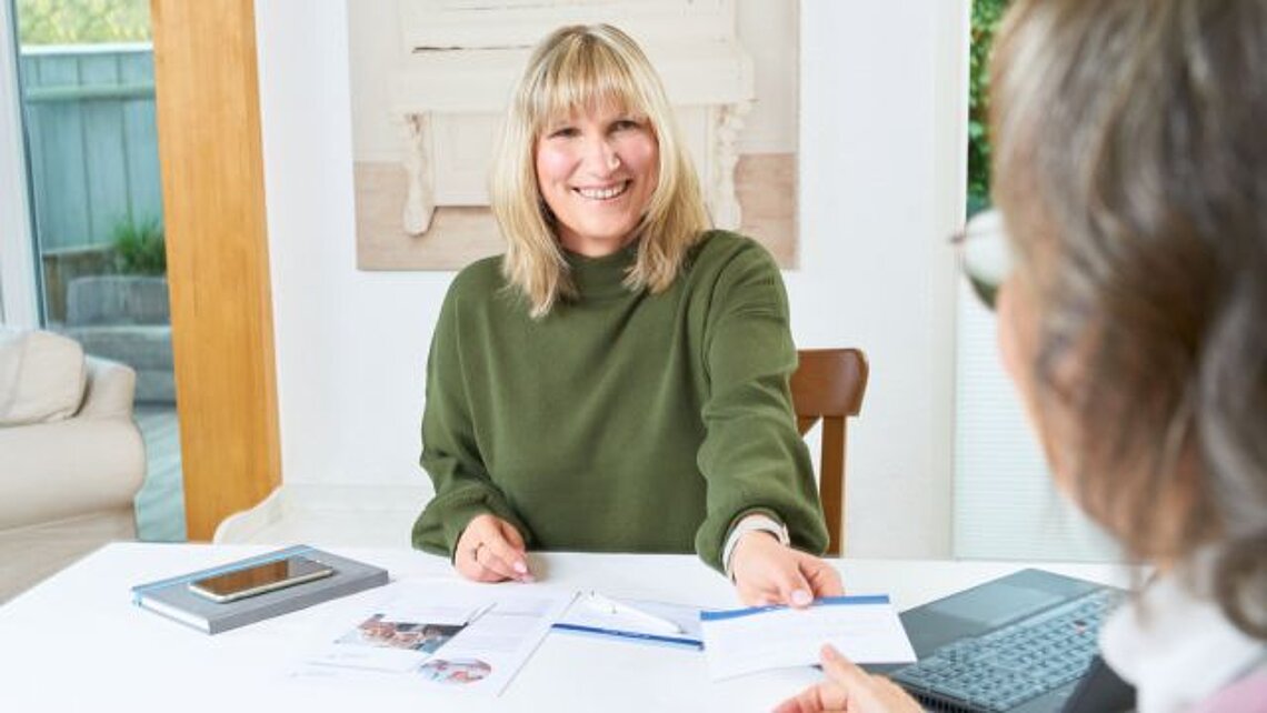 Eine Pflegeberaterin sitzt lächelnd am Tisch einer Klientin und bietet Infomaterial an.