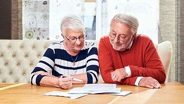 Ältere Frau und älterer Mann sitzen an einem Tisch und lesen konzentriert gemeinsam, um sich zur Pflege von Angehörigen zu informieren.