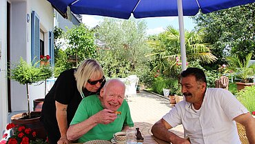 Drei Menschen auf einer Terrasse unter einem blauen Sonnenschirm. In der Mitte ein älterer Mann.