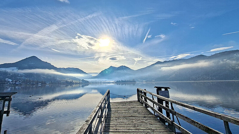 Ein Steg, der in einen großen See führt. Hinter dem See liegen mehrere Berge. Sie spiegeln sich, ebenso wie der blaue Himmel, der von einzelnen Wölkchen durchzogen ist, im Wasser. 