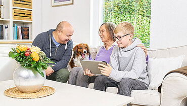 Drei Generationen sitzen gemeinsam auf einer Couch und nehmen über das Tablet eine Beratung per Videogespräch in Anspruch. Der Familienhund ist auch dabei.