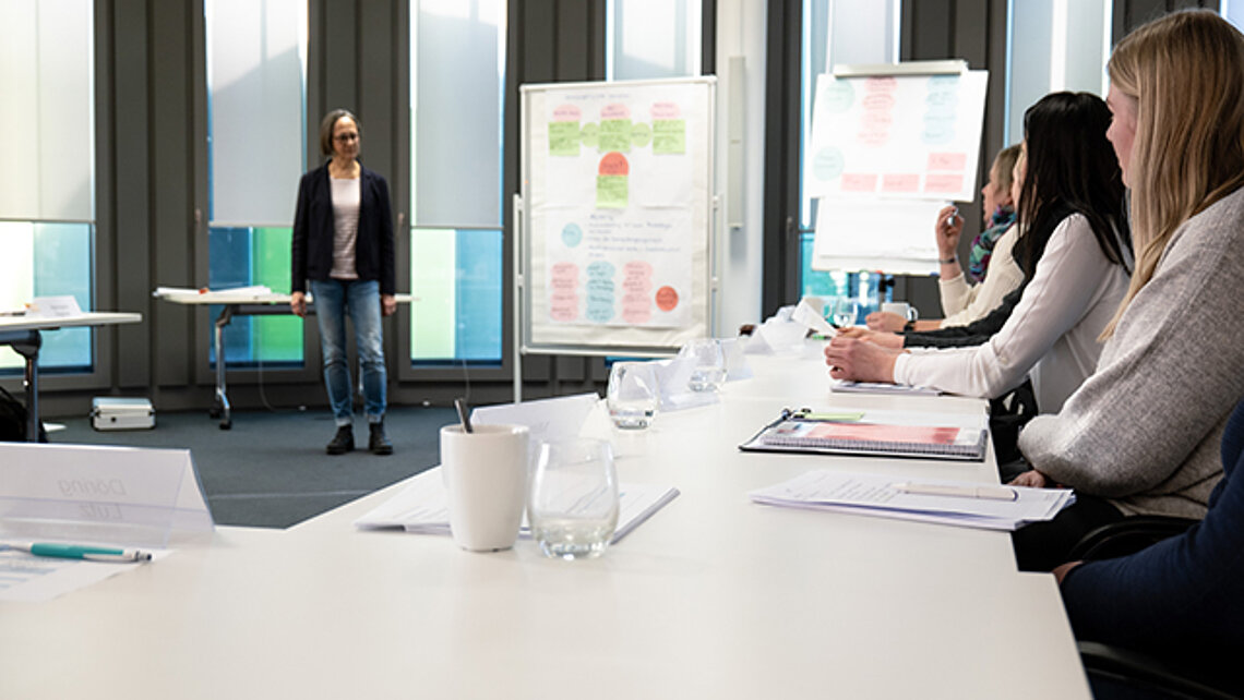 Eine Dozierende steht vorne im Raum. Neben ihr stehen Flipcharts. An der Seite des Bildes sitzen Personen an Tischen und folgen dem Unterricht.