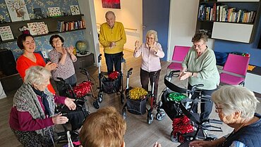 Die Rollatortanzgruppe der Tagespflege am Weidenbusch ist zu sehen. Die älteren Damen und Herren stehen mit den Rollatoren in einem Kreis und klatschen in die Hände. Auf den Rollatoren liegen Pompoms.