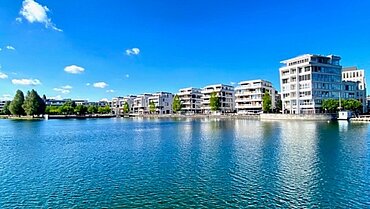 Ein See bei blauem Himmel mit Häusern im Hintergrund.