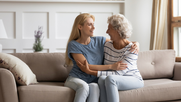 Zwei Frauen, die rechte älter als die linke, sitzen zusammen auf der Couch, haben sich im Arm und lächeln sich an.