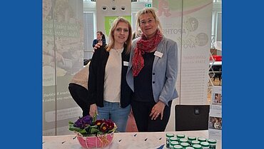 Mandy Böttger und Simone Schumacher stehen an einem Messestand Arm in Arm und lächeln in die Kamera.  