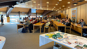 Viele Menschen sitzen an Tischen und schauen nach vorne, wo eine Frau steht und in ein Mikrofon spricht.