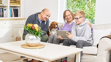 Eine Familie sitzt auf dem Sofa. Oma, Vater, Kind und Hund schauen zusammen auf ein Tablet.