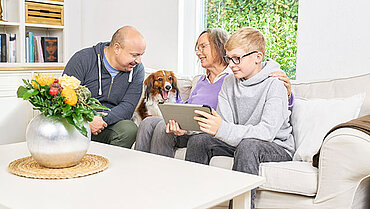 Im Bild sieht man drei Familienmitglieder gemeinsam auf dem Sofa, die an einem Tablet den Online-Pflegekurs machen.