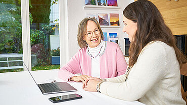 Zwei Frauen sitzen sich zugewandt nebeneinander an einem Tisch. Die ältere der beiden lächelt die jüngere an. Vor ihnen auf dem Tisch steht ein aufgeklappter Laptop, daneben liegt ein Mobiltelefon.
