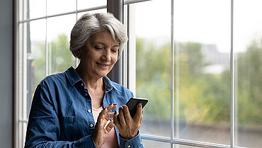 Eine Frau mittleren Alters steht lächelnd an einem Fenster und macht eine Eingabe auf ihrem Smartphone.