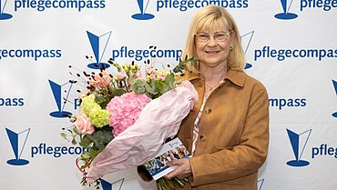 Eine blonde Frau hält einen bunten Blumenstrauß in der Hand. Im Hintergrund eine Wand mit pflegecompass-Logos.
