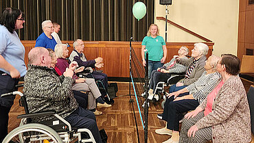 Mehrere ältere Menschen sitzen in einer Sporthalle auf zwei Seiten, getrennt von einem Netz und halten einen Luftballon in der Höhe.  
