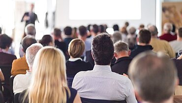 Mehrere Menschen sitzen in einem Raum und blicken in Richtung einer vortragenden Person.