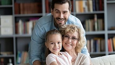 Eine Familie (Vater, Oma und Tochter) lachen vor einem Bücherregal in die Kamera.