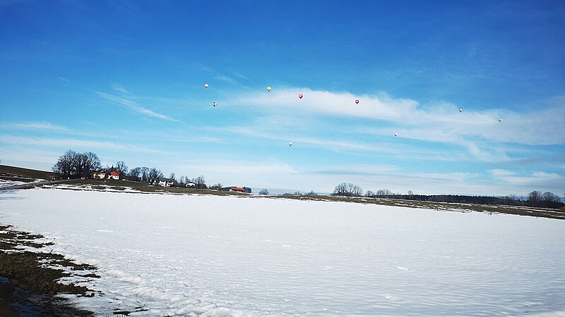 Schneebedeckte, flache Landschaft unter einem blauen Himmel. Mehrere bunte Heißluftballons fliegen.