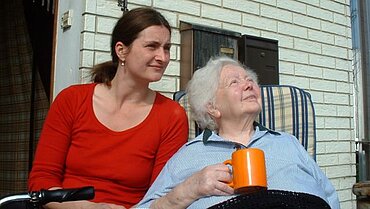 Eine ältere und eine jüngere Frau sitzen vor einem Haus in der Sonne. Die ältere Frau hat eine Tasse in der Hand. Am linken unteren Bildrand ist der Griff eines Rollators im Bildausschnitt.