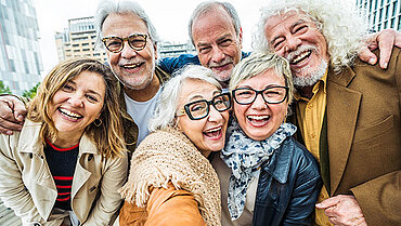 Selfie von drei lachenden Frauen und drei lachenden Männern, alle über 60 Jahre alt, draußen, vor städtischem Panorama in warmen Jacken.