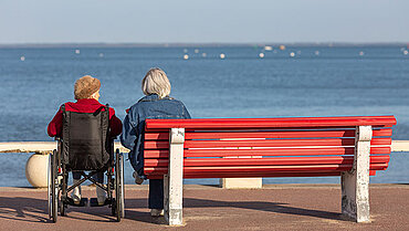 Zwei Personen schauen nebeneinander aufs Meer, die eine im Rollstuhl, die andere auf einer roten Bank sitzend.