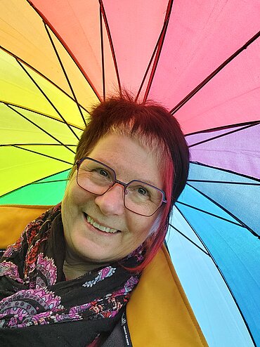 Gabi Hug vor einem aufgespannten Regenschirm in Regenbogenfarben.