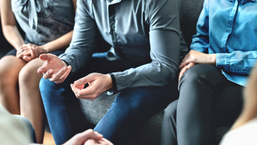 Mehrere Personen sitzen zusammen auf einer Couchbank. Die Person im Zentrum des Bildes gestikuliert.