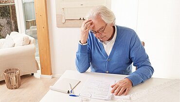 Ein Mann im höheren Lebensalter sitzt an einem Tisch und fasst sich mit seiner rechten Hand an die Stirn, vor ihm liegen Formulare und ein Notizblock mit Stift.